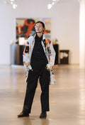 A man wearing ModaMichelle's unisex Splatter Print Car Coat over a black shirt and black trousers stands in a spacious, brightly lit art gallery or exhibition space.