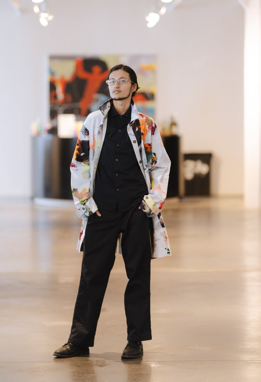 A man wearing ModaMichelle's unisex Splatter Print Car Coat over a black shirt and black trousers stands in a spacious, brightly lit art gallery or exhibition space.